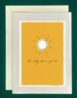 Close-up of a white greeting card on top of a cream-colored envelope, all on a forest green background. Card features a large yellow rectangle box outlined with soft clouds. In the middle is a sketched sun and cursive writing that says, "All a baby needs is your love"