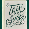 Close-up of a white greeting card on top of a cream-colored envelope, all on a forest green background. Card features the words "This Sucks" in large, fancy forest-green script