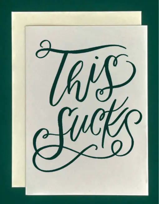 Close-up of a white greeting card on top of a cream-colored envelope, all on a forest green background. Card features the words "This Sucks" in large, fancy forest-green script