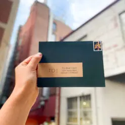 image of a stamped, forest green envelope held up in front of blurry brick buildings. The envelope has a brown, kraft paper label that reads 