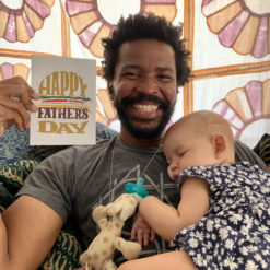 A smiling Black man holds up a Father's Day card to show the camerawhile a sleeping baby lies on his chest.
