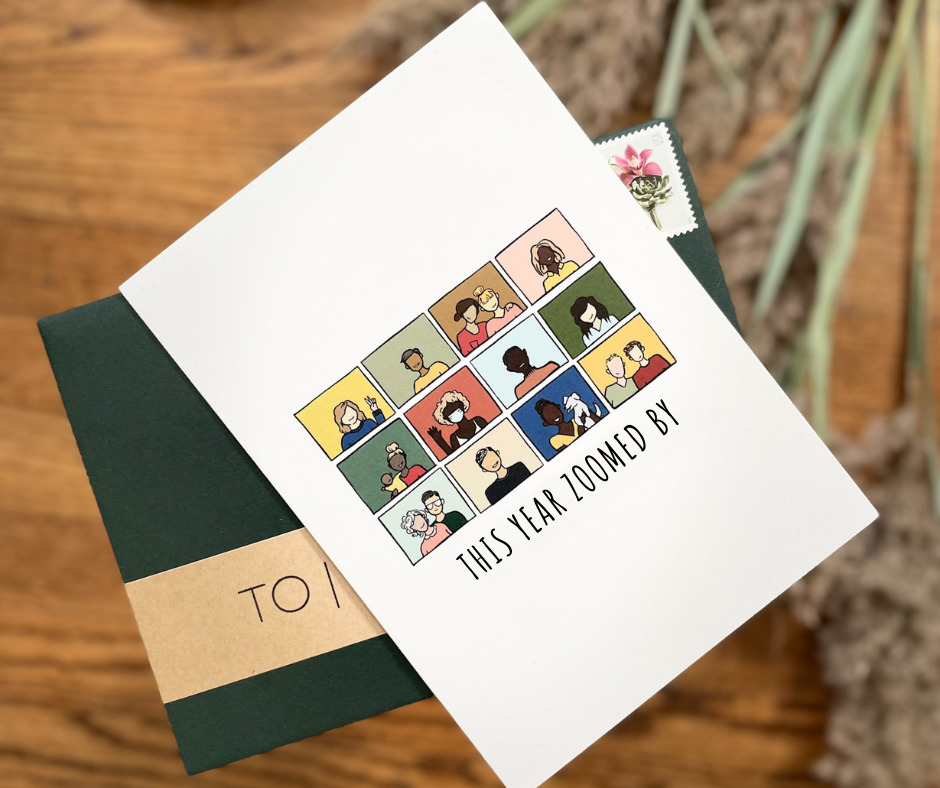 image of a white greeting card lying on a stamped, forest green envelope, all atop a blurred background of wood grain and pampas grass. the greeting card features a rectangular image comprised of 12, multi-colored rectangles in a 4x3 grid. The rectangles feature a cartoon person or people of different ages, races, ethnicities, and family structures. The text underneath reads "THIS YEAR ZOOMED BY" in a thin, handwritten font