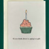 Close-up of a white greeting card on top of a cream-colored envelope, all on a forest green background. The card has a drawing of a cupcake covered in pink icing lit with a single birthday candle. The text underneath reads "If you think about it, aging is a gift."