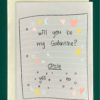 Close-up of a white greeting card on top of a cream-colored envelope, all on a forest background. Card features a handwritten note design with the words "Will you be my galentine, circle yes or no"