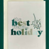 Close-up of a white greeting card on top of a cream-colored envelope, all on a forest background. The card features a sprig of holly and the words, "the best way to holiday"
