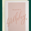 Close-up of a white greeting card on top of a cream-colored envelope, all on a forest green background. Card says happy birthday in pink script superimposed on a light pink background