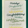Close-up of a white greeting card on top of a cream-colored envelope, all on a forest background. The card reads "Congratulations!" in many languages, all in different shades of green