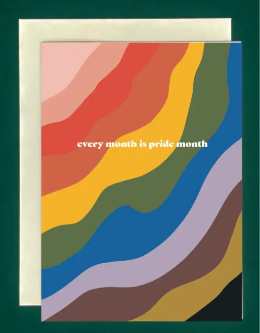 Close-up of a greeting card on top of a cream-colored envelope, all on a forest background. The greeting card is rainbow-colored, and reads, "every month is pride month"