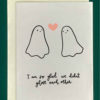 Close-up of a white greeting card on top of a cream-colored envelope, all on a forest background. Card features two simple gosht drawings, looking at one another and smiling. There is a pink heart between them. Underneath reads the text, "I'm so glad we didn't ghost each other"
