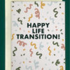 Close-up of a white greeting card on top of a cream-colored envelope, all on a forest background. The card has confetti art all over. In the center of the card are the words, "Happy Life Transition!"
