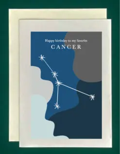 Close-up of a white greeting card on top of a cream-colored envelope, all on a forest background. The card features the constellation Cancer superimposed on abstract blobs of different shades of blues. Text at the top of the greeting card reads, "Happy Birthday to my favorite Cancer"