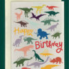 close up product image of a white greeting card on a cream envelope, all on a forest colorerd background. The card is filled with various dinosaurs, all differently-colored. In the middle are the words "Happy Birthday" in a warm handwritting