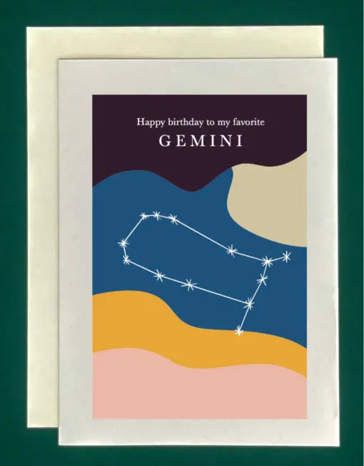 Close-up of a white greeting card on top of a cream-colored envelope, all on a forest background. The card features the constellation Gemini superimposed on abstract blobs of different colors - dark blue, dark purple, goldenrod, and pink. Text at the top of the greeting card reads, "Happy Birthday to my favorite Gemini"