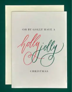 Close-up of a white greeting card on top of a cream-colored envelope, all on a forest green background. The card reads, "Oh by golly have a holly jolly christmas." The words "holly jolly" are written in large, red and green script letters.