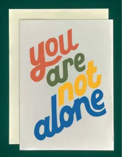 Close up of a white greeting card on top of a cream-colored envelope, all on a forest background. Greeting card reads "you are not alone." each word is a different color - red, green, yellow, blue