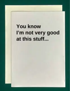 Close-up of a white greeting card on top of a cream-colored envelope, all on a forest background. The card reads, "You know I'm not very good at this stuff..." in black text