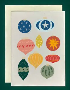 Close-up of a white greeting card on top of a cream-colored envelope, all on a forest background. Card art features a collage of multi-colored ornaments