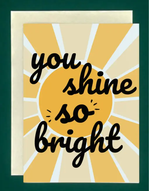 image of a greeting card that reads "you shine so bright" in bold, black cursive font in front of a geometric sun on a light blue background