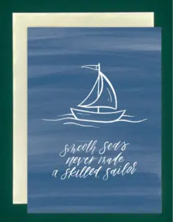 Close-up of a royal blue greeting card on top of a cream-colored envelope, all on a forest green background. Card with a blue painted background and a drawing of a sailboat with words that read "smooth seas never made a skilled sailor" in script
