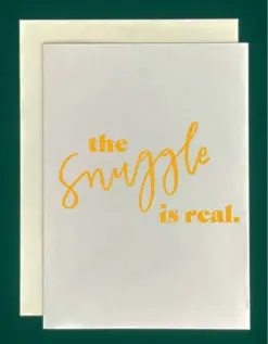 Close-up of a white greeting card on top of a cream-colored envelope, all on a forest background. The card reads, "The snuggle is real" in golden yellow text