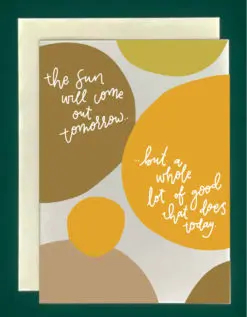 Close up of a white greeting card on top of a cream-colored envelope, all on a forest background. The card features different sized circles in various shades of golden and taupe. The card reads, "The sun will come out tomorrow... but a whole lot of good that does today."