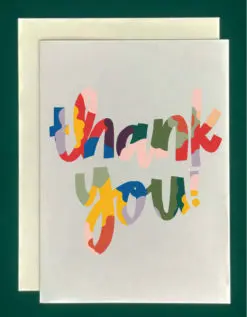 Close-up of a white greeting card on top of a cream-colored envelope, all on a forest green background. The card reads, "Thank You" in colorful script hand lettering