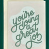 Close-up of a white greeting card on top of a cream-colored envelope, all on a forest background. Card reads, "You're doing a great job" in green script, superimposed on a lighter green, abstract blob
