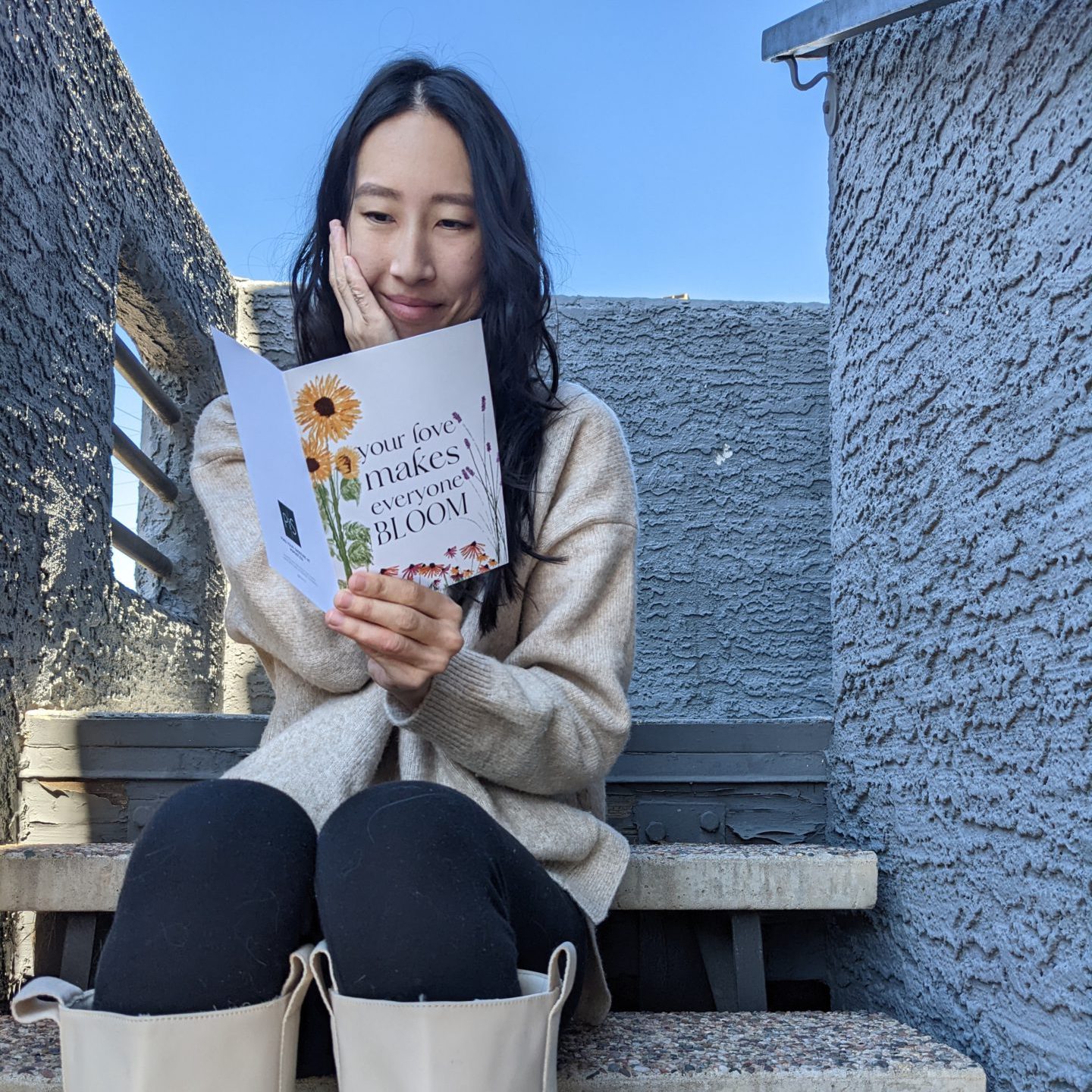 A woman is sitting on outside steps and smiling with one hand on her face while reading a greeting card that she is holding. The greeting card has flowers on the front and the words, "Your love makes everyone bloom"