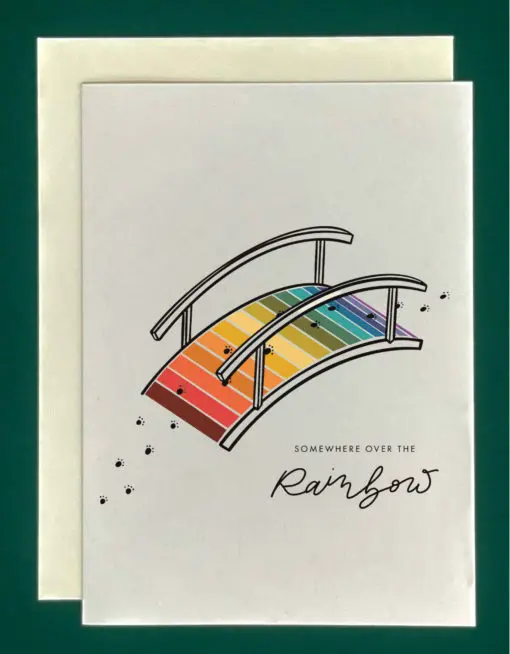 Product Image of a Rainbow Bridge greeting card. The card features a colored drawing of a bridge. Each plank of the bridge is a different color, and the colors together make up a rainbow. A trail of paw prints walks over the bridge. Written on the front are the words "Somewhere over the rainbow."